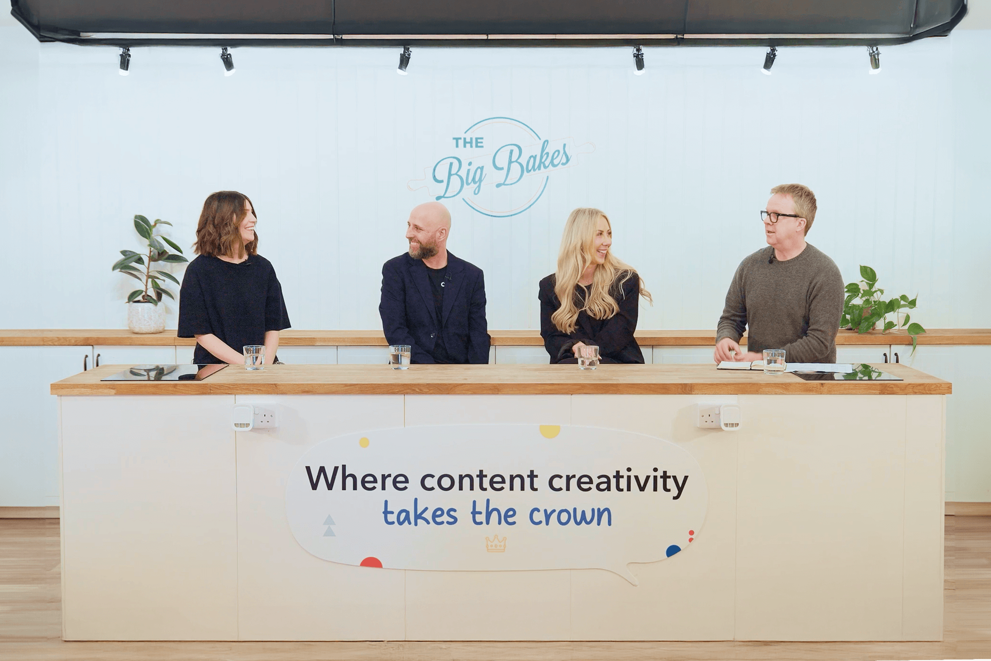 Four team members seated at a desk with "The Big Bakes" logo and text "Where content creativity takes the crown" on wall behind them