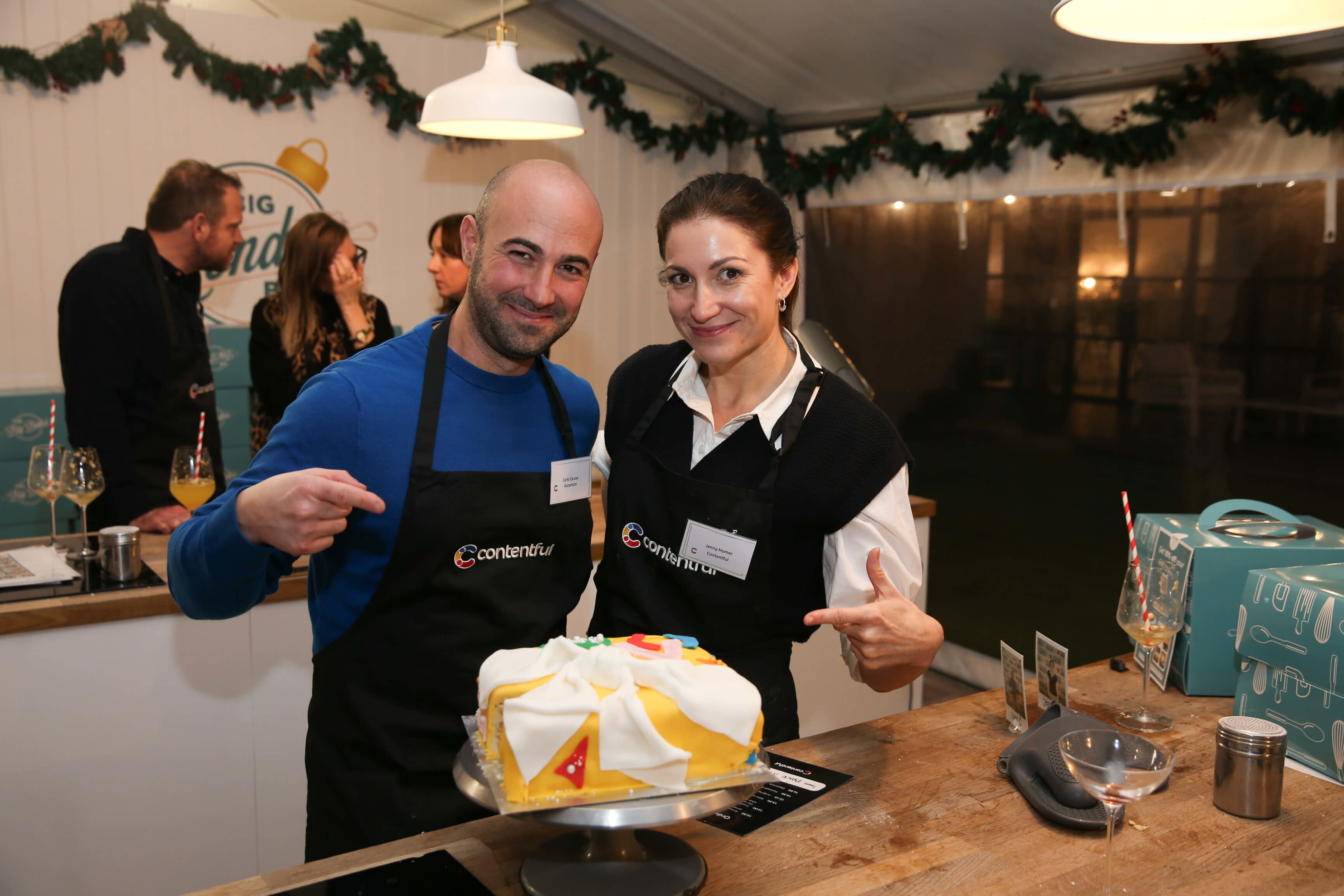 Two Contentful team members in black aprons present a decorated white and yellow cake at a holiday event with evergreen garlands